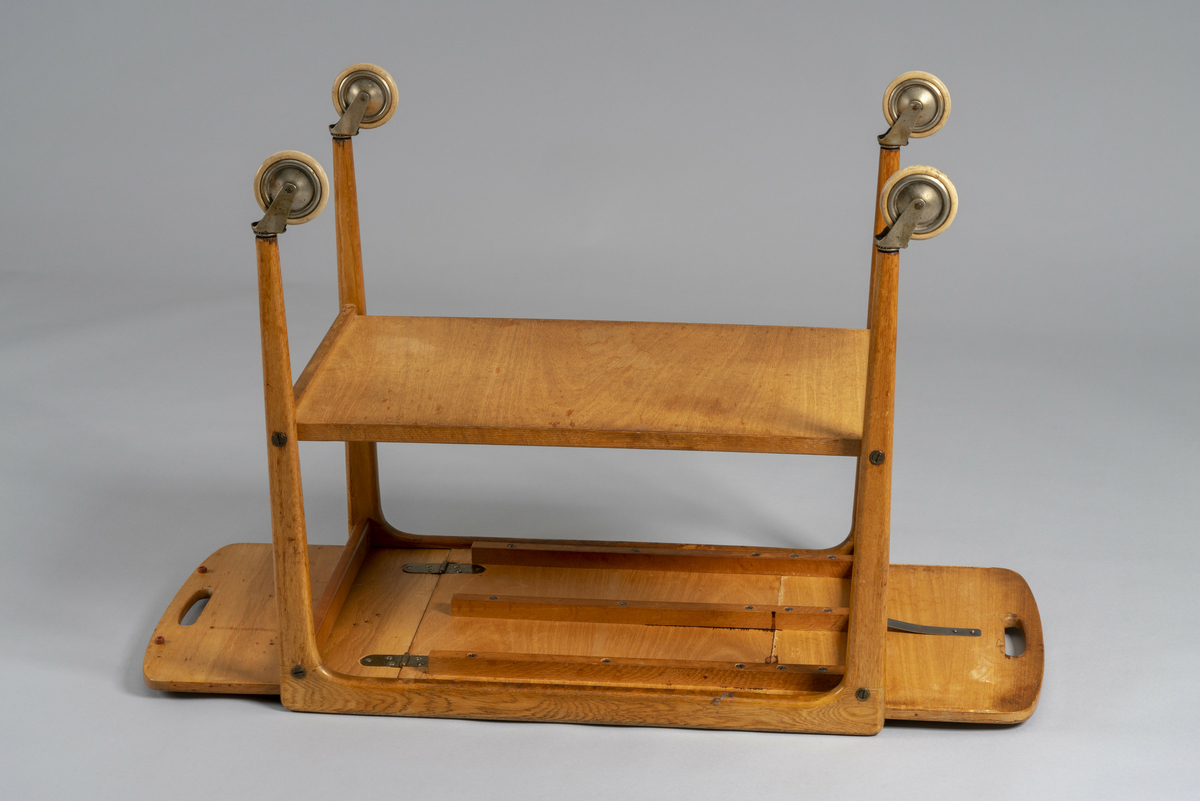 Trillebord med to klaffer. Topplaten kan skyves sidelengs slik at en eller begge klaffer er slått opp. Posisjonen låses med et metallbeslag under. Hull til håndtak i begge kortender av bordplaten, hylleplate under og fire hjul.  Har tilhørt Sara Fabricius.
