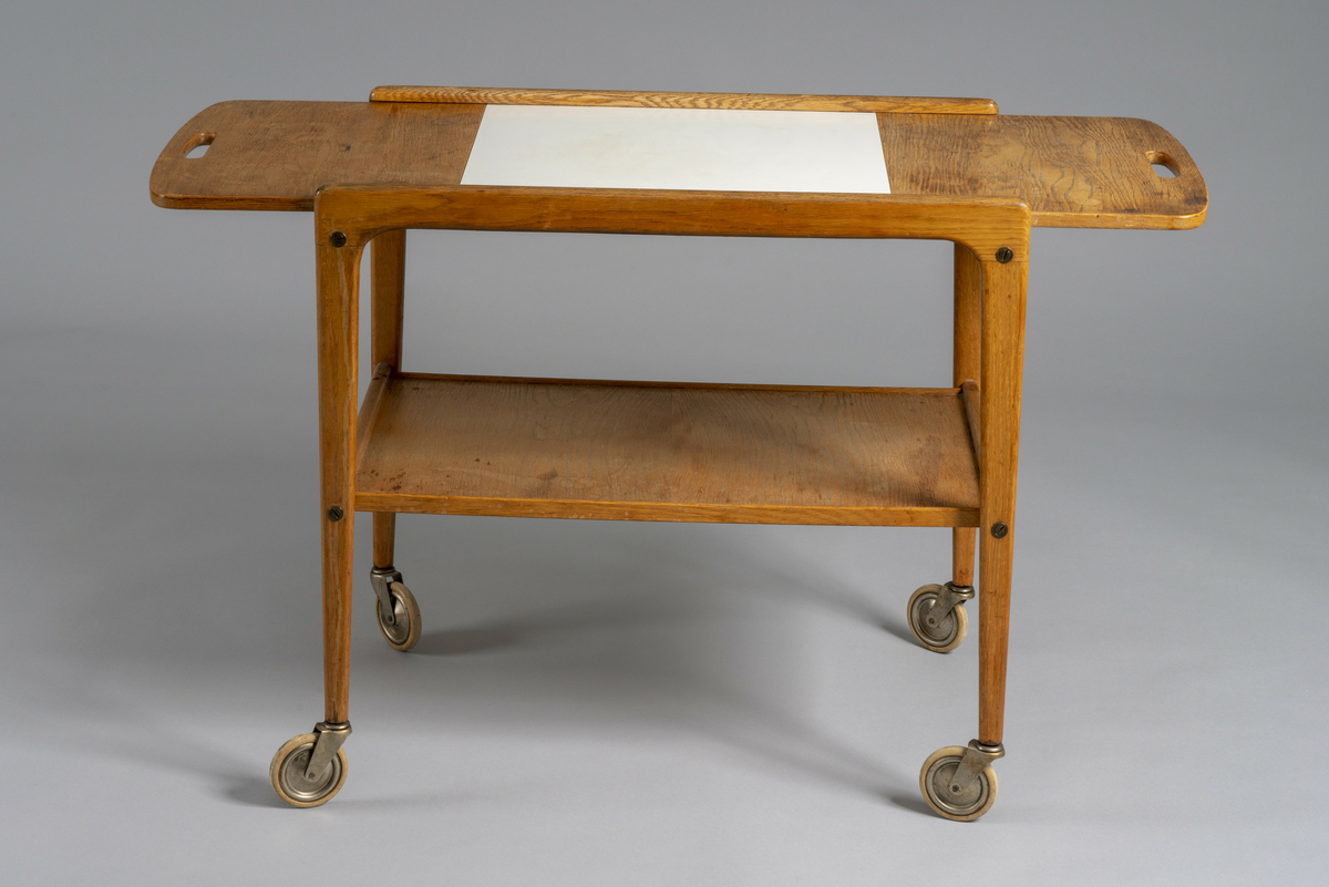 Trillebord med to klaffer. Topplaten kan skyves sidelengs slik at en eller begge klaffer er slått opp. Posisjonen låses med et metallbeslag under. Hull til håndtak i begge kortender av bordplaten, hylleplate under og fire hjul.  Har tilhørt Sara Fabricius.