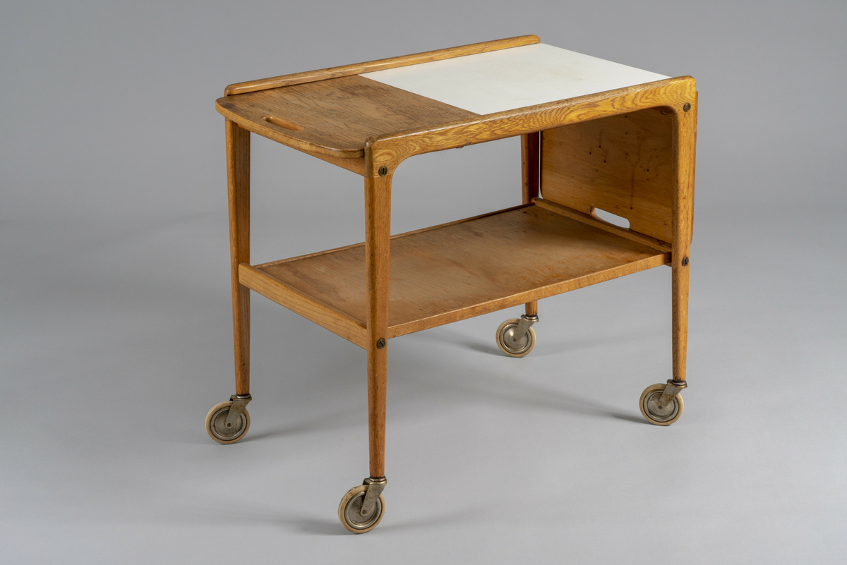Trillebord med to klaffer. Topplaten kan skyves sidelengs slik at en eller begge klaffer er slått opp. Posisjonen låses med et metallbeslag under. Hull til håndtak i begge kortender av bordplaten, hylleplate under og fire hjul.  Har tilhørt Sara Fabricius.