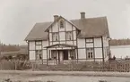 Ett större bostadshus med veranda, på landet. I bakgrunden syns en sjö. Ca 1910.