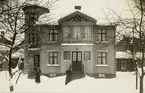 Ett bostadshus i snö, med två kvinnor stående i trädgården framför. Ca 1914.