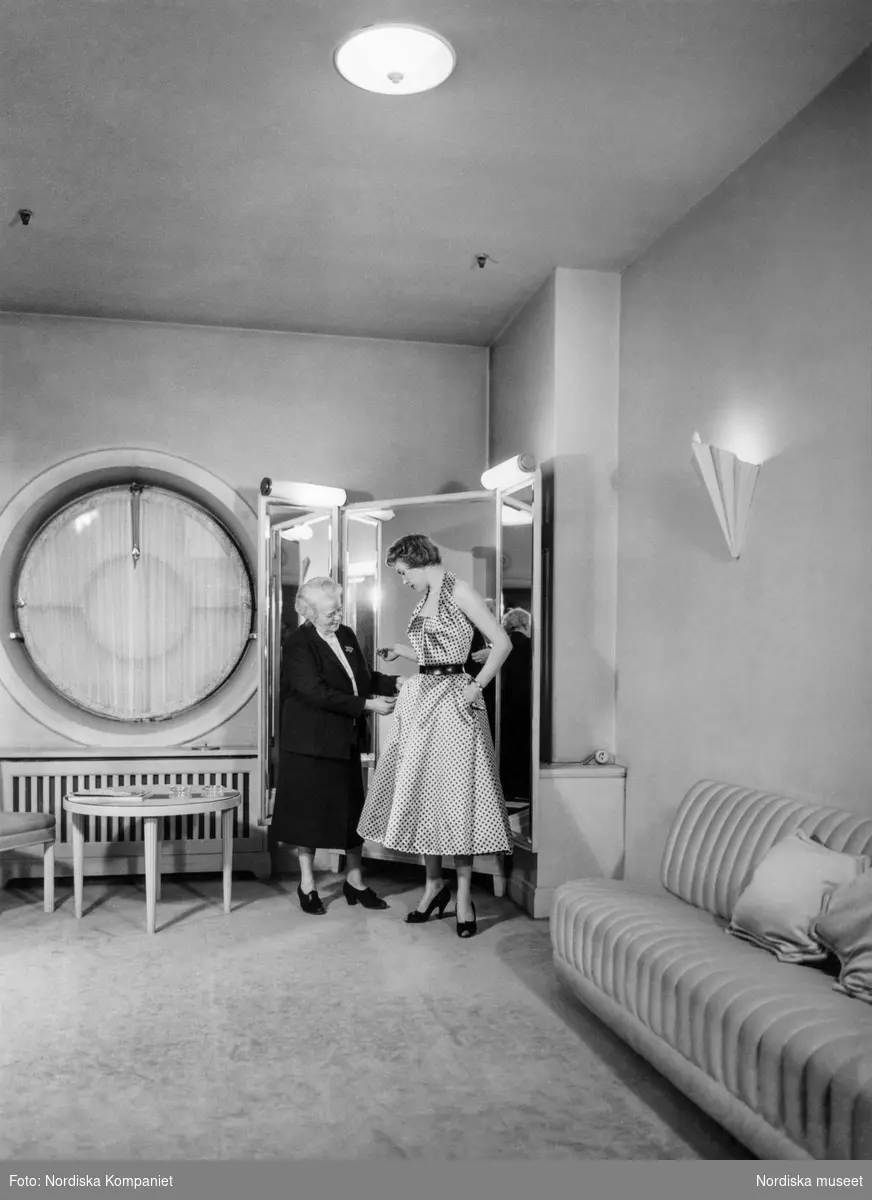 Kvinna provar prickig klänning i provrummet vid NK:s Franska damskrädderi. Hon assisteras av äldre kvinna i mörk dräkt; proverskan Greta Nisshammar (1892-1982) en av ateljéns trotjänare. När hon gick i pension 1965 hade hon tjänstgjort i drygt 40 år.