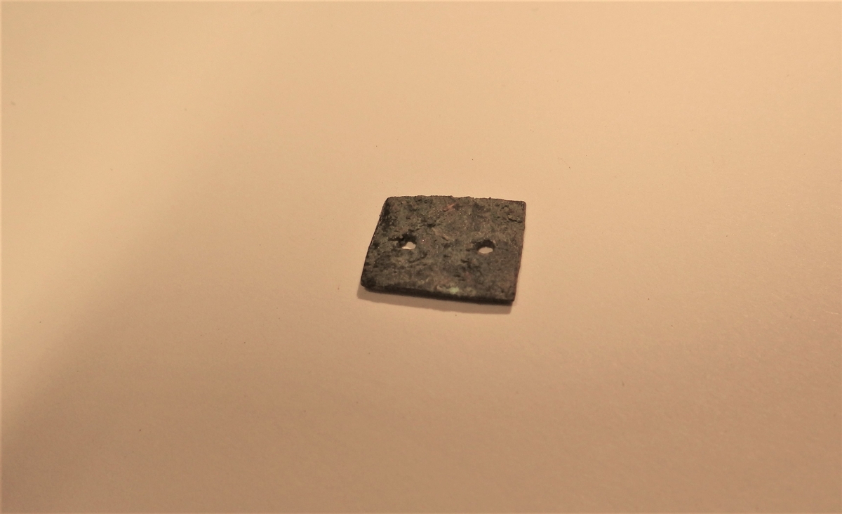 MÅL: L. 1, 5 CM,  B. 1, 1 CM, TILNÆRMET REKTANGULÆRT STYKKE AV TYNN BRONSEPLATE. TO SMÅ SYMETRISKE PLASSERTE HULL.