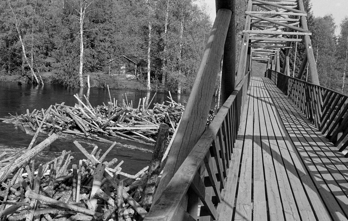 Tømmerhauger ved to av betongkarene som bærer Norsk Skogbruksmuseums (nå Norsk skogmuseum) bru over Klokkerfossen i Glomma, Elverum i Hedmark. Fotografiet er tatt under fløtinga i 1982. Egentlig skulle et helst ikke gå tømmer i denne delen av elva. Fløterne brukte nemlig å legge ei ledelense fra den østre elvebredden ved Elverum prestegard mot et steinskjær sentralt i elveløpet ovenfor det stedet der elva delte seg i to. Fra dette punktet ville strømdraget i elva vanligvis trekke fløtingsvirket mot Prestfossen, på den nordvestre sida av Prestøya. Årsaken til at Prestfossen var foretrukket som fløtingsled framfor Klokkerfossen var dels at vannføringa og dybden i Prestfossen var større, dels at brua over denne delen av elveløpet var ei hengebru, uten brukar som kunne fange fløtingstømmeret. Den Klokkerfossbrua vi ser på dette fotografiet ble bygd vinteren 1963-64 i såkalt tømmersprengverkskonstruksjon med 20 meters spenn mellom brukarene, og med en samlet lengde på 108 meter. Den ble erstattet av ei ny bru bygd med samme type bærekonstruksjon i 2005.