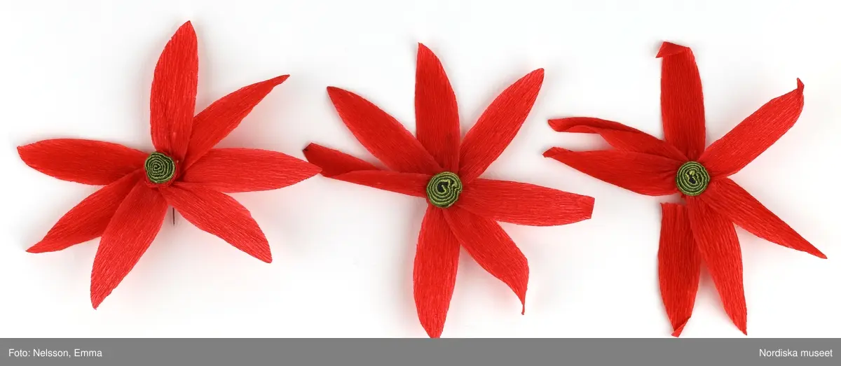 a-l) Tolv stycken hemtillverkade julgransprydnader av rött kräpp-papper i form av blommor/julstjärnor, med upphängningsanordning av knappnålar. 

Lena Kättström Höök 2019-03-21