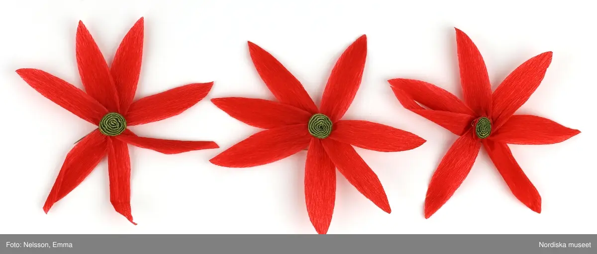 a-l) Tolv stycken hemtillverkade julgransprydnader av rött kräpp-papper i form av blommor/julstjärnor, med upphängningsanordning av knappnålar. 

Lena Kättström Höök 2019-03-21