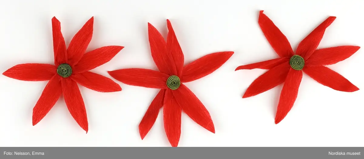 a-l) Tolv stycken hemtillverkade julgransprydnader av rött kräpp-papper i form av blommor/julstjärnor, med upphängningsanordning av knappnålar. 

Lena Kättström Höök 2019-03-21