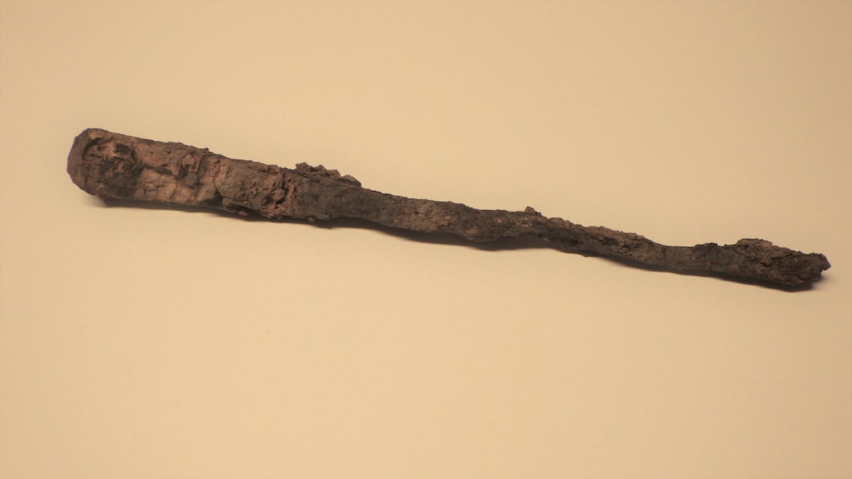 MALV: L. 16 CM B. (SKAFT) 1, 5 CM, LAGET AV EN JERNSTANG, ANT. OPPR. REKTANGULÆRT TVERR-SNITT. STERKT KORRODERT. RESTER AV SKAFT BEVART. DET ER BREDEST ØVERSTHVOR DET OGSÅ ER AVRUNDET, OG SMALNER NEDOVER MOT SELVE REDSKAPET. SERUT SOM SKAFTET ER LAGET AV TYNNE JERNPLATER, HOLDT SAMMEN MED NAGLERAV KOBBERLEG. HULROM UNDER PLATENE, FYLT MED NOE GULHVITT KALKAKTIG. 