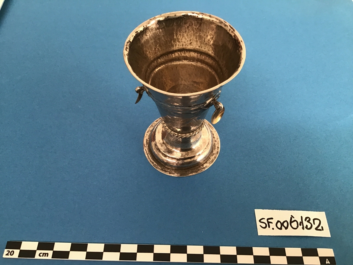 Sølvbeger med stett. Konisk form med utsvinga munning og profilert stett. 1/4 over stetten ein tauflettingskrans kring staupet. Ca 3/4 oppe på korpus heng tre gullforgylte lauv.
Inngravert girlander motiv med Z-strek.
Stetten ser ut til å vere hamra