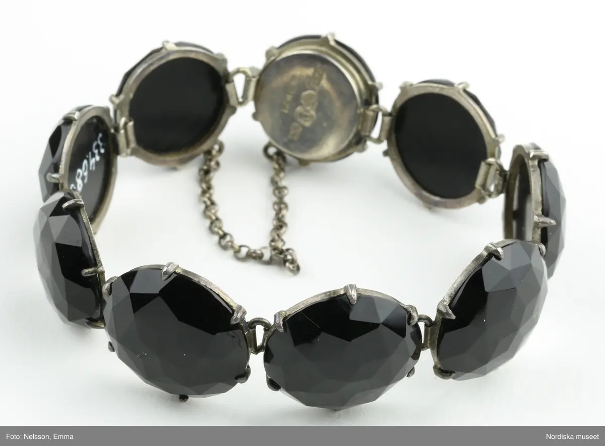 Armband av onyx och silver. Nio stycken runda fasettslipade onyxstenar infattade i silver. Fäste mellan två stenar med silvertunga som skjuts in i fäste (lösgörs genom att trycka ned metallpigg). Kedja mellan stenarna vid öppningen. Stämplar i silvret under en av stenarna vid fästet "CWT"/ kattfot (tre kronor)  / St Erik / M6 / Hedblom.".
/Leif Wallin 2021-09-24