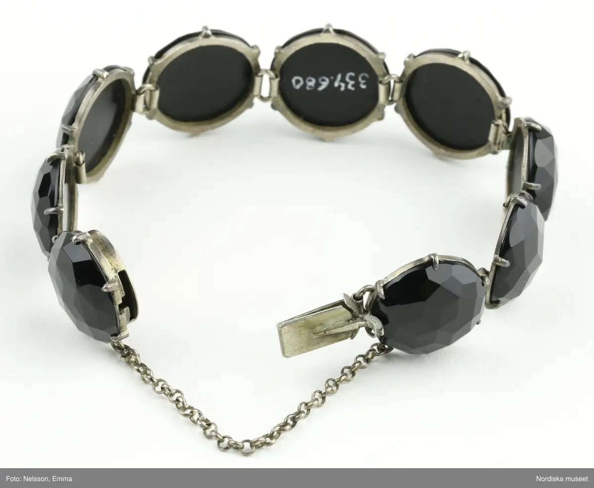Armband av onyx och silver. Nio stycken runda fasettslipade onyxstenar infattade i silver. Fäste mellan två stenar med silvertunga som skjuts in i fäste (lösgörs genom att trycka ned metallpigg). Kedja mellan stenarna vid öppningen. Stämplar i silvret under en av stenarna vid fästet "CWT"/ kattfot (tre kronor)  / St Erik / M6 / Hedblom.".
/Leif Wallin 2021-09-24