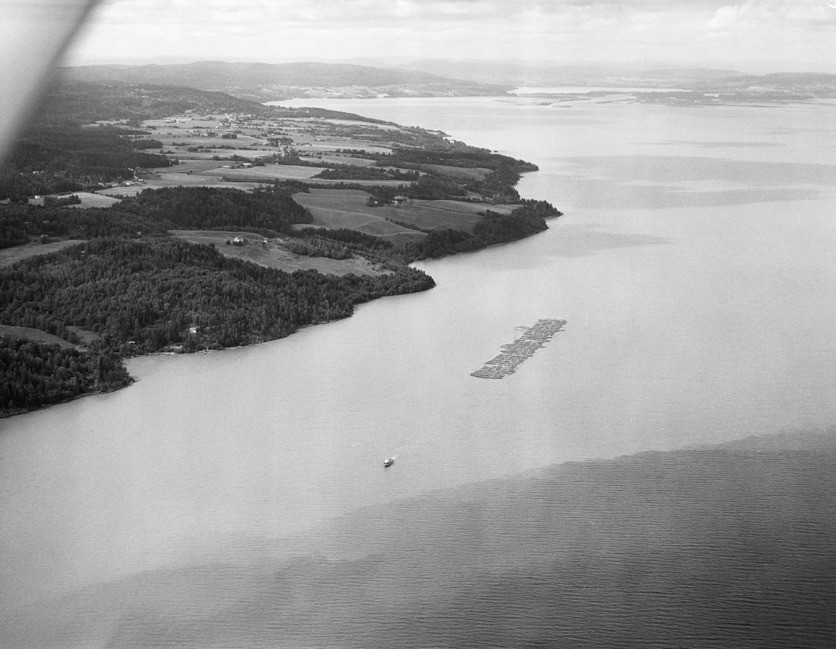 Tømmerslep over innsjøen Øyeren sommeren 1985.  Tømmer fra Østerdalen og Solør, Gudbrandsdalen, Mjøstraktene og bygdene på Øvre Romerike ble fløtet løst ned til Fetsund lenser i nordenden av denne innsjøen.  Her ble virket fra gammelt av sortert og «soppet» eller «moset» (buntet), og det som skulle til Lillestrøm, Strømmen og Oslo ble skilt fra det som skulle videre nedover vassdraget.  Da dette fotografiet ble tatt var det bare storbedriften Borregaard fabrikker i Sarpsborg som fortsatt fløtet, så noe særlig sortering var det ikke snakk om lenger.  Tømmeret ble moset i høvelige bunter med hver sin «grime» (vaierbindsel).  Inntil 200 av disse mosene ble bundet sammen i et slep som ble buksert forsiktig ned gjennom en meandrerende djupål i det grunne deltapartiet lengst nord i innsjøen.  Når et slikt slep kom fram til Sofiedal ved Engelsvika på innsjøens vestside, like ved grensa mellom Rælingen og Enebakk kommuner, ble slepet ankret opp og slepebåten Mørkfos gikk tilbake til Fetsund for å hente nok et slep av samme type.  Når også dette var framme ved Sofiedal ble de to slepene koplet sammen, slik at Mørkfos kunne trekke begge to sørover med ei lang trosse.  I noen tilfeller ble det også hektet på bommer med løstømmer fra «etterrensken» langs sjøen.  Slepet kom gjerne fram til Sleppetangen, ved nedre Glommas utløp fra Øyeren, seint på kvelden.  Da ble slepet oppankret mot land, slik at lokale fløtere påfølgende dag kunne løsne vaierbindslene og la tømmeret flyte løst nedover mot tømmerrennene forbi kraftverksdammene nedover i vassdraget.  Mørkfos gikk nordover mot Fetsund igjen om natta, slik at båten kunne fortsette slepearabeidet derfra neste morgen.  Dette flyfotografiet er tatt etter at to slep var koplet sammen til ett, altså nedenfor Sofiedal, idet det slepes langs Øyerens vestbredd.  Opptaket ble gjort i 1985, det siste året det foregikk kommersiell tømmerfløting i dette vassdraget.