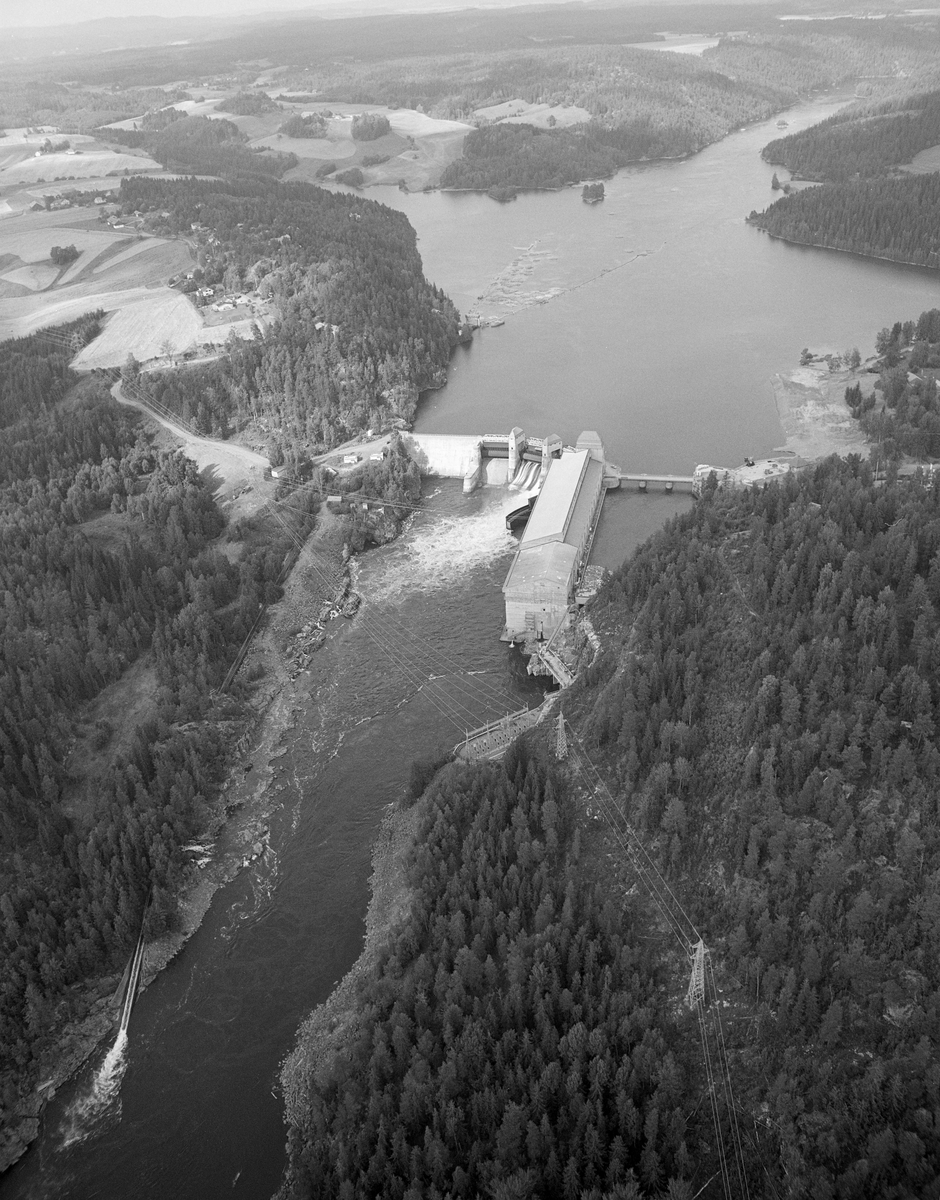 Flyfotografi tatt over Mørkfoss/Solbergfoss kraftstasjon i Askim, sett fra en posisjon over den østre elvebredden. Her var Glomma grense mellom Askim og Spydeberg kommuner. Bildet er tatt ned mot elva med kraftstasjonen og kraftverksdammen. Dette kraftverket ble bygd i perioden 1917-1924 med staten (som eide ovenforliggende Mørkfoss med 10,5 meters fall) og Oslo kommune (som eide mellomliggende Vittenbergfoss, Skråperudfoss og Halfredsfoss, som hadde 5 meters fall ved lavvann). Dammen ble plassert på terskelen i den opprinnelig todelte Solbergfossen, som inntil da hadde hatt cirka 2,5 meters fallhøyde ved lavvann.  Etter at det ble reist en massiv betongdam ved Solbergfossen kunne man heve vannspeilet cirka 25 meter, på nivå med den ovenforliggende innsjøen Øyeren, som dermed ble en del av reservoaret til dette kraftverket. For å kunne gjøre det store inngrepet i vassdraget som dette kraftverksprosjektet innebar, måtte utbyggerne forhandle med Fredrikstad Tømmerdirektion, som hadde hevd på å bruke Nedre Glomma som transportåre for tømmer. På dette tidspunktet var det vanligvis 6-7 millioner stokker årlig som passerte fossen på veg mot sagbruk og papirfabrikker i området ved Fredrikstad og Sarpsborg. Forhandlingene om hvordan dette skulle løses involverte tung ingeniørfaglig og juridisk ekspertise. Det gikk halvannet tiår før man endte med en tunnelløsning som begge parter var fornøyde med. Kraftstasjonen ble tegnet av den kjente arkitekten Bredo Greve (1871-1931). Da kraftstasjonen ble satt i drift (1924) hadde den 7 Francis-turbiner. I løpet av 1930-åra ble det installert ytterligere tre. Det vi ser i forgrunnen på dette fotografiet er dammen, som hadde tre overløpsåpninger med fagverksbruer over. Inntaksbassenget befant seg i det området som på dette bildet ligger bak kraftverksbygningen. Fløtingstømmeret ble ført forbi kraftverket via ei renne på nordvestsida av vassdraget. Vi skimter påstikkingslensa på kraftverksdammens vannspeil og rennas innløp i elva nedenfor dammen på den nedre delen av dette bildet. Fløtinga i Glomma ble avviklet etter 1985-sesongen. Produksjonskapasiteten ved anlegget er seinere betydelig utvidet - jfr. fanen «Opplysninger».