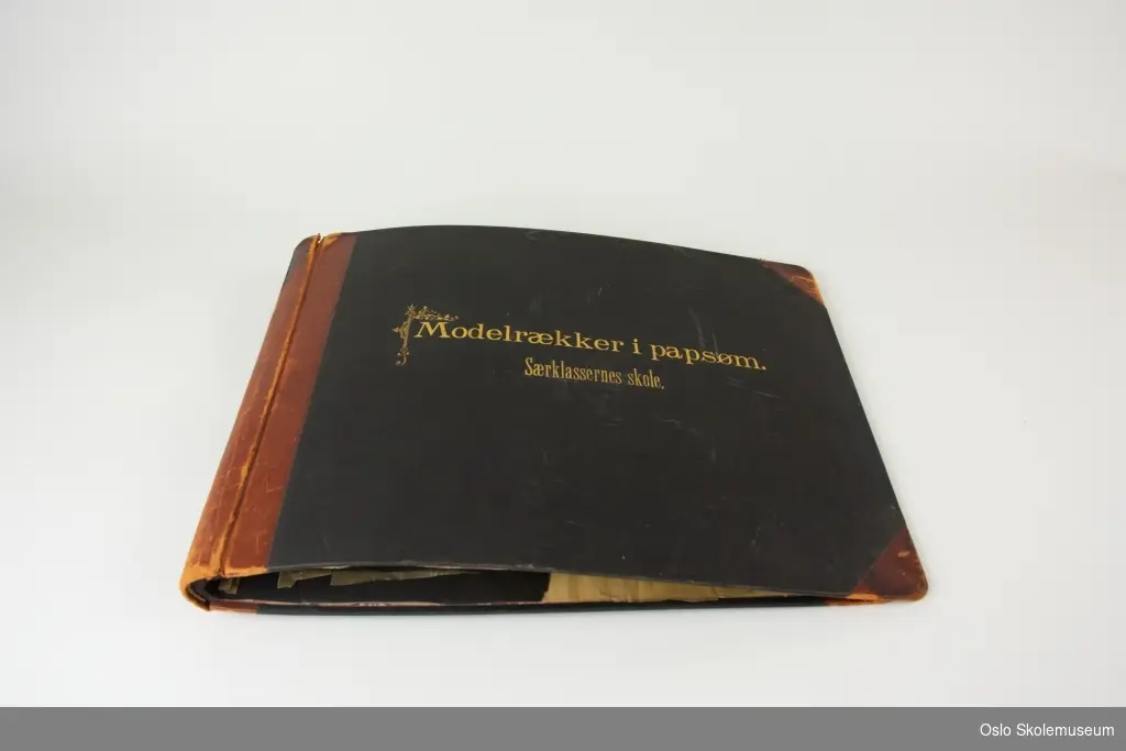 Svart og brunt album med pappsøm. Albumet har en svart perm med rygg og hjørner i brunt skinn samt en tekst i gull. Inne i albumet er det svarte sider med pappsøm. Alle disse har løsnet fra permen.