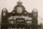 Äreport av trä och granris med bl a Gustaf V:s monogram utanför järnvägsstationen i Växjö, vid kungligt besök 1913. 
Många nyfikna har samlats i korsningen Kungsgatan/Norra Järnvägsgatan.