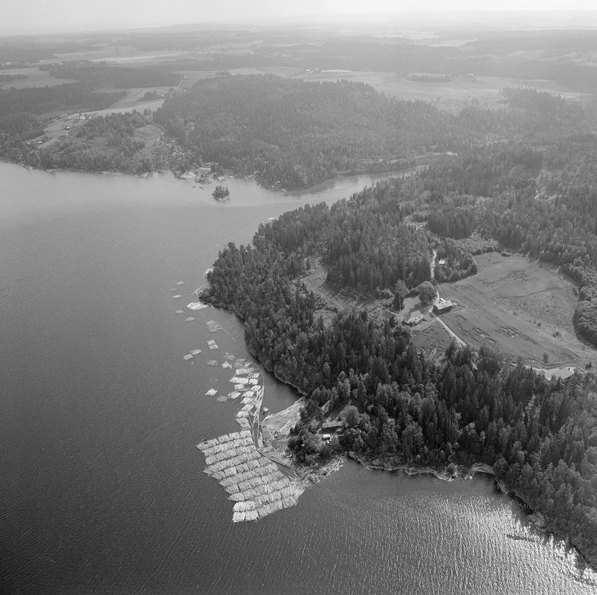 Flyfotografi tatt over Sleppetangen i søndre del av innsjøen Øyeren, hvor det var oppankringssted for tømmerslep fra Fetsund lenser i nordenden av sjøen.  På vannspeilet ser vi et oppankret slep, samt en del «moser» (tømmerbunter) som driver langs land nedover mot elveutløpet ved Mørkfoss.  Vanligvis ble også «grimene» (vaierbindslene) rundt mosene tatt av ved Sleppetangen, men når det var vestavind kunne løstømmer bli drevet tilbake mot innsjøen i stedet for mot elva.  Derfor hendte det, som på dette fotografiet, at mosene ble sendt hele nedover mot tømmerrenna ved kraftverket Solbergfoss, hvor grimene ble tatt av.  Vi ser innløpet til den nedre delen av Glomma ved Mørkfoss på øvre del av bildeflata.  På høydedraget vest for oppankringsstedet Sleppetangen ligger gardsbruket Sandvik omgitt av åkrer og skog.  Til høyre, nederst på bildeflata, speiles sollyset i vika Bogen, bakenfor Bogneset. 