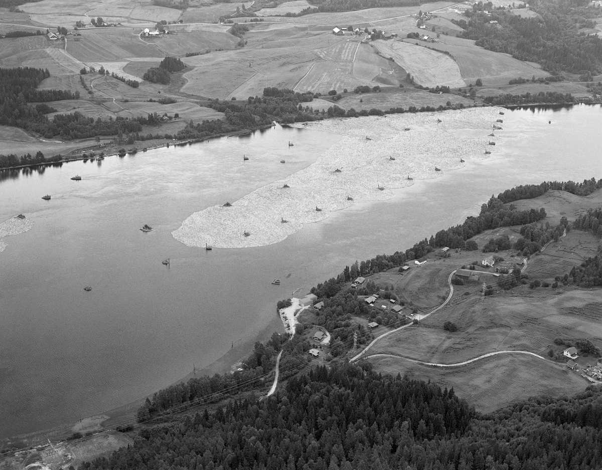 Flyfotografi, tatt ned mot Bingen lenser i Glomma, Sørum, Akershus. Denne kunstruksjonen skulle samle opp og holde på løstømmer som kom flytende fra ovenforliggende distrikter. Fra gammelt av lå sorteringslensa der tømmer som skulle til sagbrukene på Nedre Romerike ble skilt fra det som skulle videre mot brukene i Nedre Glomma. Fra 1861, da den nye sorteringslensa nedenfor jernbanebrua ved Fetsund ble satt i drift, ble anlegget ved Bingen ombygd til et reservoar, der tømmeret ble samlet for deretter å bli sluppet i høvelige dagsrasjoner videre nedover til sorteringsanlegget 6-7 kilometer lengre nede ved elveløpet. Utover sommeren og høsten kunne tømmerbeholdningen i attholdslensa ved Bingen være på flere millioner stokker. Anlegget hadde 51 lensekar - tømmerkister som var fylt med stein - som holdt lensa og tømmeret på plass. Fotografiet er tatt i motstrøms retning, fra en posisjon over den østre elvebredden, like sør for kommunesenteret Sørumsand. Kar. Tømmerkar.