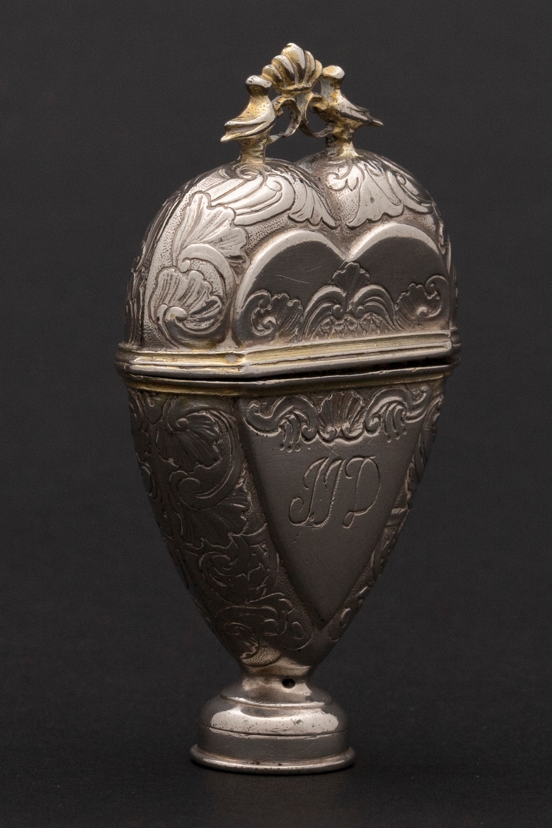 Hjerteformet luktevannshus i sølv med rund fot og ornament mellom to duer på lokket. Innvendig lokk med gjennombrutt bladornament. Korpus er dekorert med rokokko-ornamenter. Graverte initialer på forsiden, årstall på baksiden og speilmonogram under fotstykket. Innvendig forgylt.