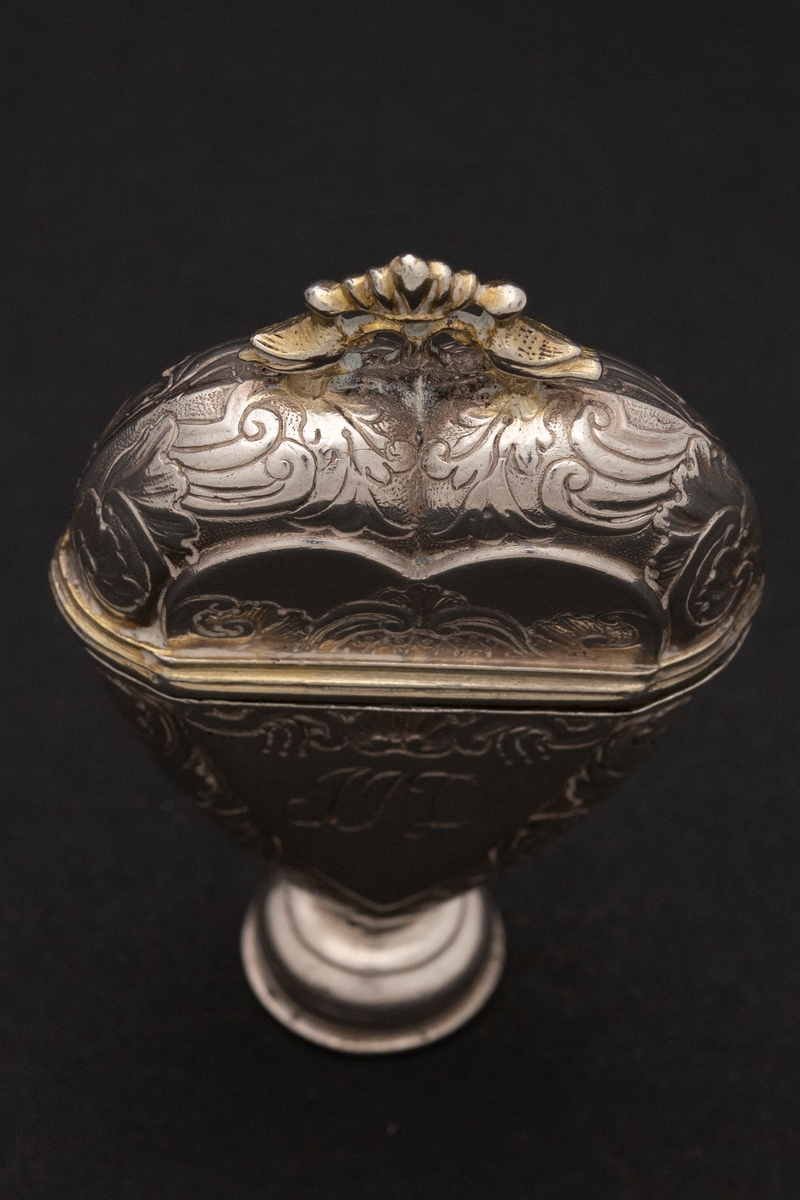 Hjerteformet luktevannshus i sølv med rund fot og ornament mellom to duer på lokket. Innvendig lokk med gjennombrutt bladornament. Korpus er dekorert med rokokko-ornamenter. Graverte initialer på forsiden, årstall på baksiden og speilmonogram under fotstykket. Innvendig forgylt.