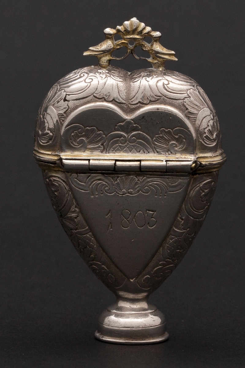 Hjerteformet luktevannshus i sølv med rund fot og ornament mellom to duer på lokket. Innvendig lokk med gjennombrutt bladornament. Korpus er dekorert med rokokko-ornamenter. Graverte initialer på forsiden, årstall på baksiden og speilmonogram under fotstykket. Innvendig forgylt.