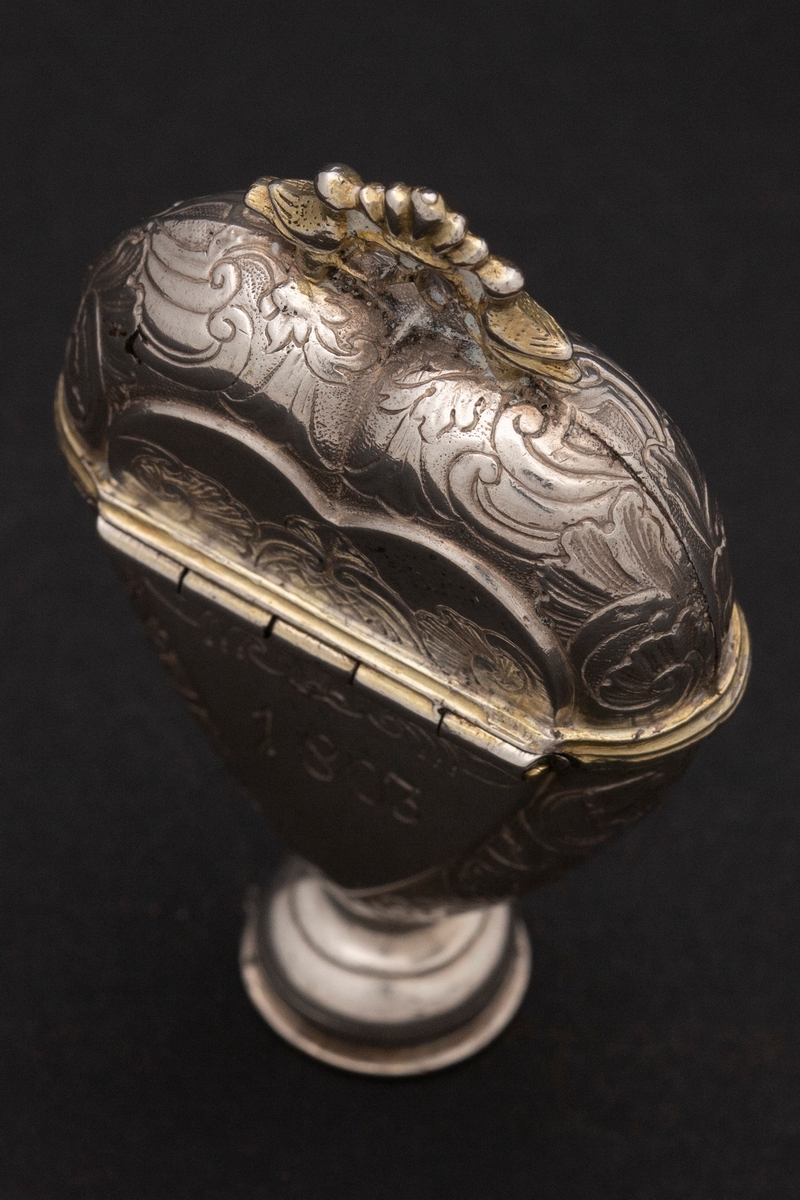 Hjerteformet luktevannshus i sølv med rund fot og ornament mellom to duer på lokket. Innvendig lokk med gjennombrutt bladornament. Korpus er dekorert med rokokko-ornamenter. Graverte initialer på forsiden, årstall på baksiden og speilmonogram under fotstykket. Innvendig forgylt.