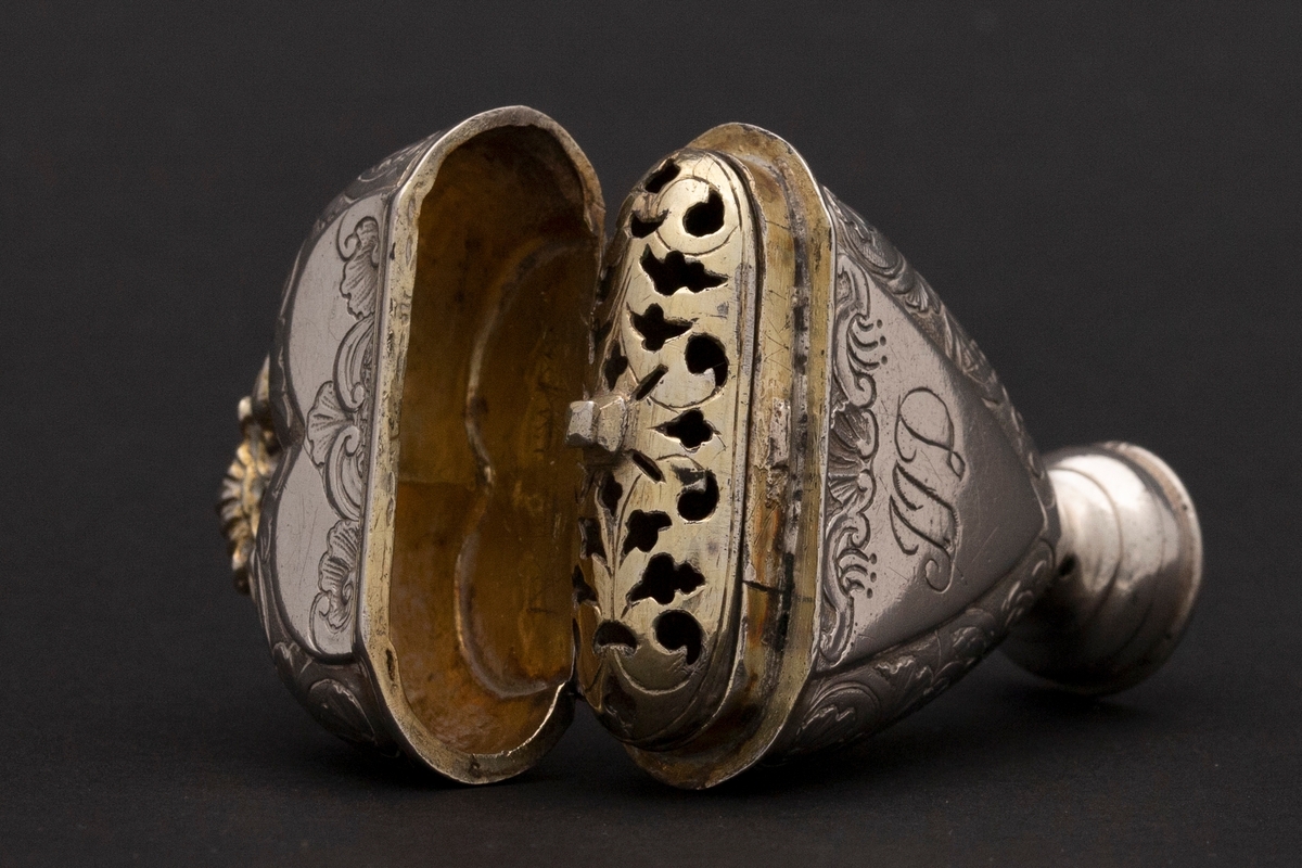 Hjerteformet luktevannshus i sølv med rund fot og ornament mellom to duer på lokket. Innvendig lokk med gjennombrutt bladornament. Korpus er dekorert med rokokko-ornamenter. Graverte initialer på forsiden, årstall på baksiden og speilmonogram under fotstykket. Innvendig forgylt.