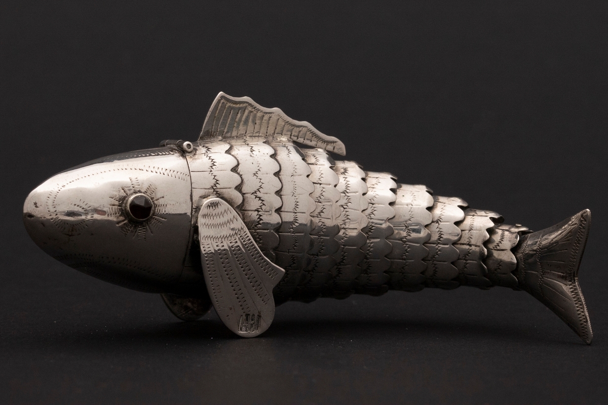 Etui i leddet fiskeform med øyne av rødt glass. Fiskens hode danner lokket. Innvedig rom. Sikksakk-dekor.