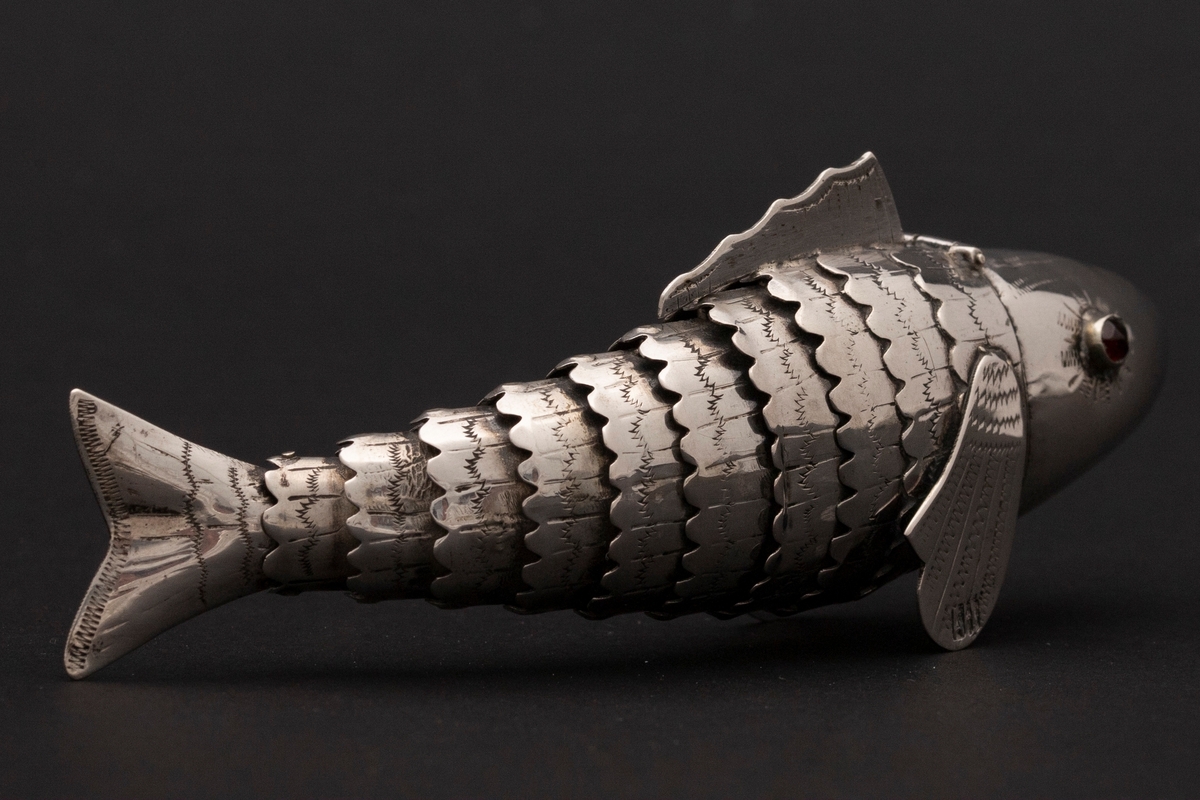 Etui i leddet fiskeform med øyne av rødt glass. Fiskens hode danner lokket. Innvedig rom. Sikksakk-dekor.