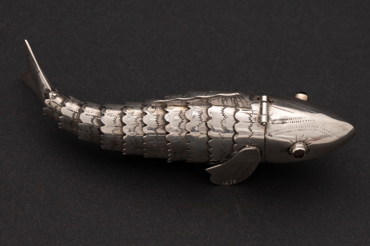 Etui i leddet fiskeform med øyne av rødt glass. Fiskens hode danner lokket. Innvedig rom. Sikksakk-dekor.