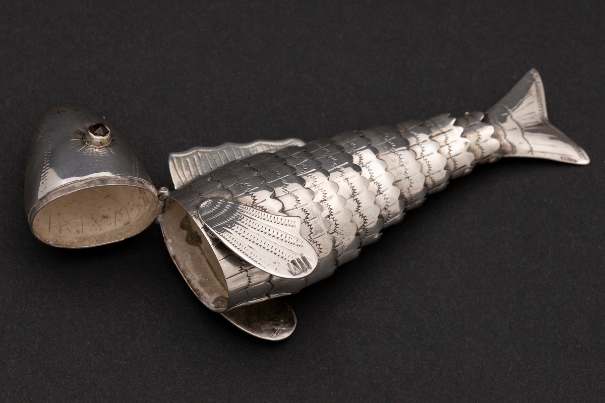 Etui i leddet fiskeform med øyne av rødt glass. Fiskens hode danner lokket. Innvedig rom. Sikksakk-dekor.