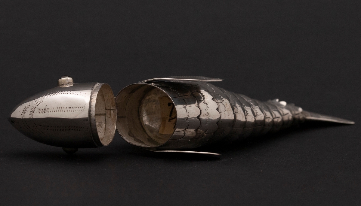 Etui i leddet fiskeform med øyne av rødt glass. Fiskens hode danner lokket. Innvedig rom. Sikksakk-dekor.