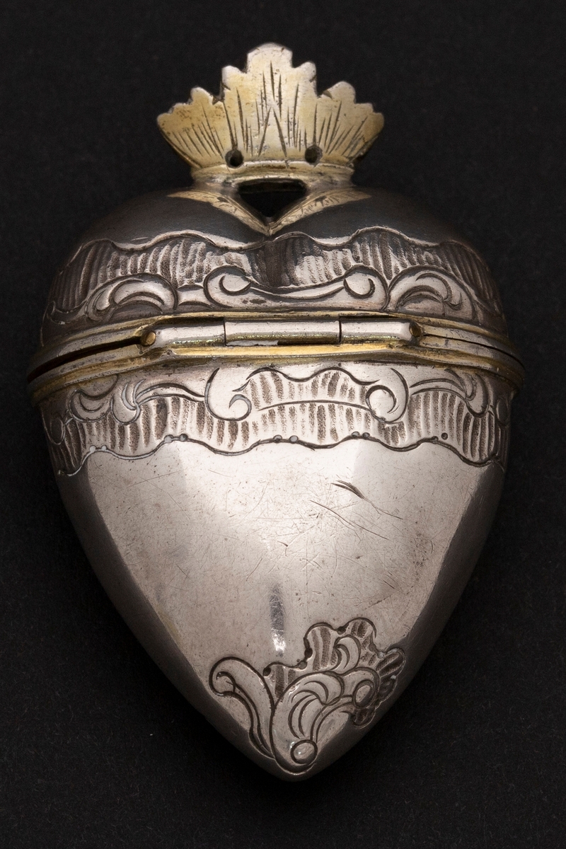 Hjerteformet luktevannshus i sølv uten fot. Delvis gjennombrutt, åpen, forgylt krone med strekgravyr på lokket. Gravert rokokko-ornamentikk på korpus; kartusj med rester av forgylling på forsiden. Forgylt kant rundt åpningen og innvendig forgylt.