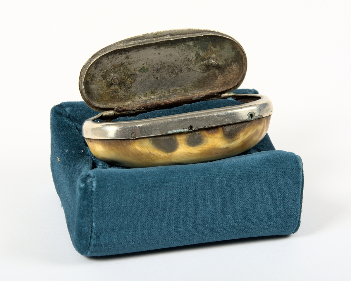 Ovalt formad snusdosa med bottendel snidad av kohorn. Lock och överdel är tillverkat i nysilver. Snusdosan vilar på en kudde av blå sammet, och snusdosan är även vadderad på insidan med blå sammet.