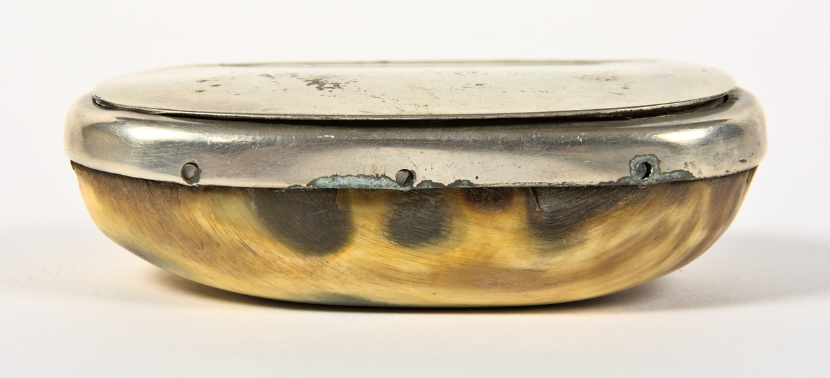 Ovalt formad snusdosa med bottendel snidad av kohorn. Lock och överdel är tillverkat i nysilver. Snusdosan vilar på en kudde av blå sammet, och snusdosan är även vadderad på insidan med blå sammet.