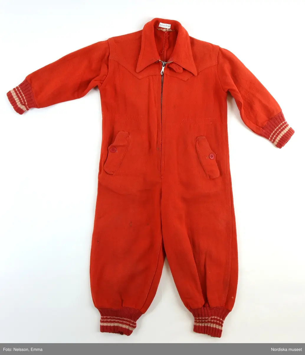 Katalogkort:
"Overall av rött kypertvävt ruggat ylletyg. 
Två framstycken, två bakstycken, fram besparing med snibbig nederkant. Knäppning mitt fram med långt blixtlås av metall (Wico), överst slejf med knapphål samt röd knapp, krage med spetsiga snibbar. Två snett iskurna fickor fram, ficklock med knapphål samt knapp, fickpåsar av blått bomullstyg. Ärm av ett stycke, i nederkant samt i benets nederkant mudd av rött ullgarn med vita ränder. Hank i nacken. 
Anm: Nött. Små fläckar på höger ben."