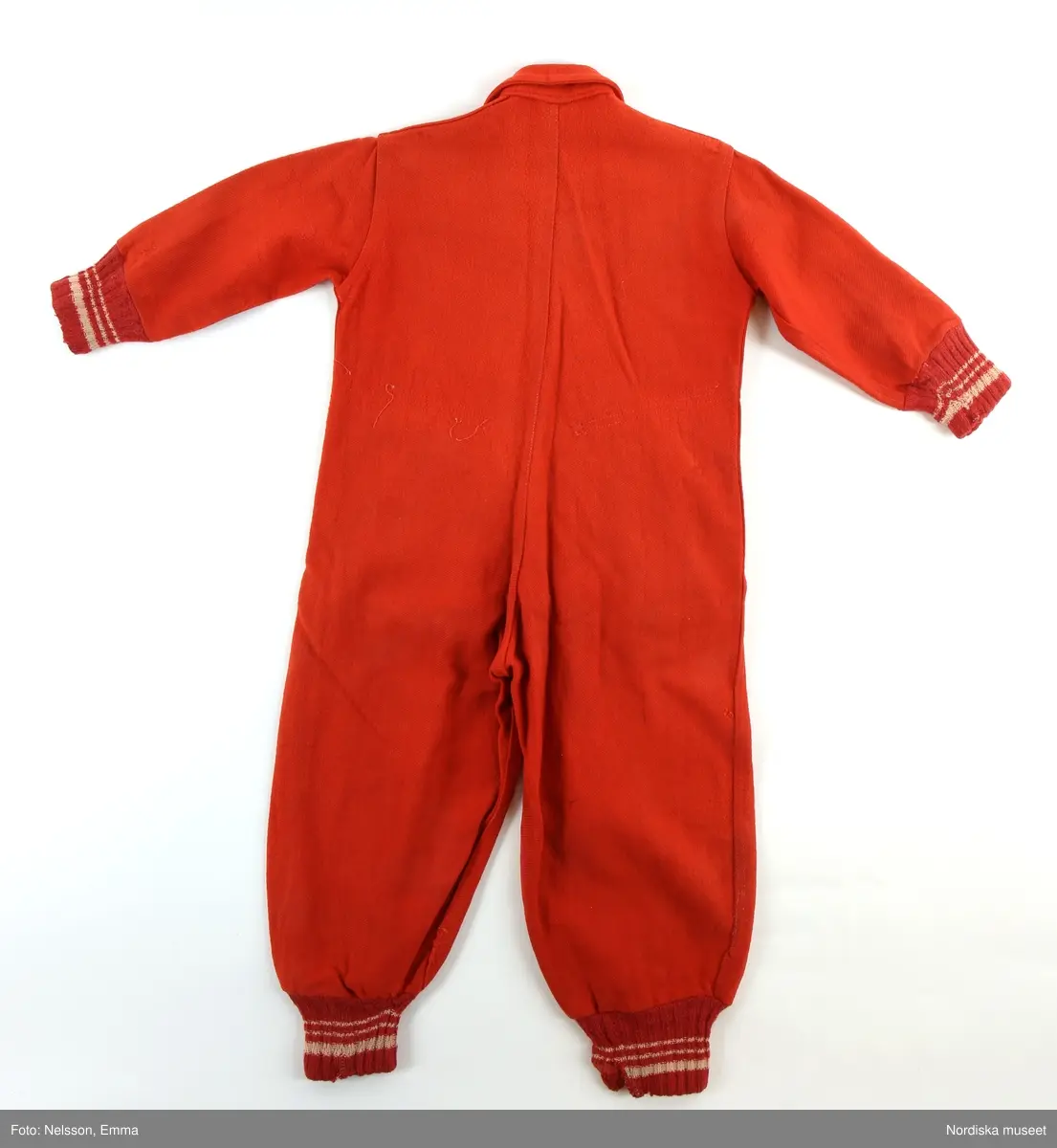 Katalogkort:
"Overall av rött kypertvävt ruggat ylletyg. 
Två framstycken, två bakstycken, fram besparing med snibbig nederkant. Knäppning mitt fram med långt blixtlås av metall (Wico), överst slejf med knapphål samt röd knapp, krage med spetsiga snibbar. Två snett iskurna fickor fram, ficklock med knapphål samt knapp, fickpåsar av blått bomullstyg. Ärm av ett stycke, i nederkant samt i benets nederkant mudd av rött ullgarn med vita ränder. Hank i nacken. 
Anm: Nött. Små fläckar på höger ben."