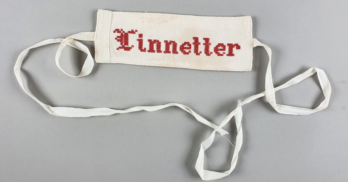 To etiketter for lintøy, med broderte bokstaver i rødt og blått. Bånd (snor) festet på midten av hver kortside.