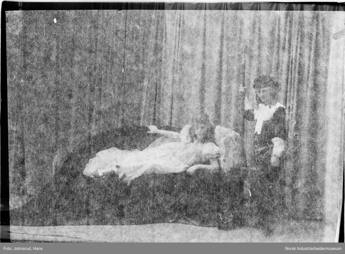 To personer på en scene, jenten til venstre ligger på en sofa i kjole med krone. Personen til høyre er stående og holder et spyd. Oppsett av teaterstykket Tornerose.
