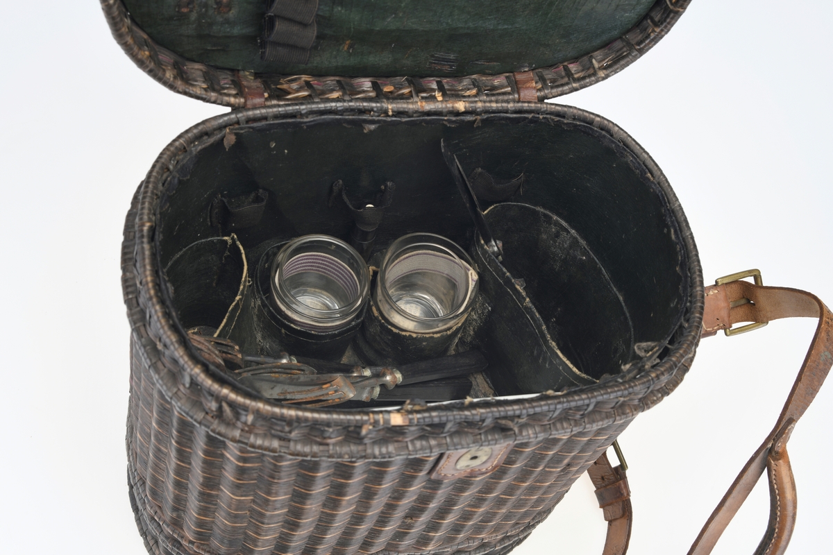 En piknikkurv med følgende innhold: 1 matboks, 2 større vannglass, 2 mindre vannglass, 4 hvite tallerkener, 4 kniver, 4 gafler og 1 beholder for salt/pepper. Kurven er flettet av pil eller lignende tre og er i ulik nyanser av brunt. Kurven er todelt, og i den nederste delen er det plass til matboksen, mens i den øverste delen det øvrige av piknikkutstyr. Beggel delene er foret med tesktil som er behandlet med granitol og deretter farget grønt. Langs kanten oppe (i begge delene) er det en svart tungekant av skinn. I den øverste delen av kurven er det festet strikk i lokket og i sidene i kurven for å feste utstyr. Det er også laget fire rom for å sette piknikkutstyr i. Rommene er laget av tekstil behandlet med granitol og farget grønn. Utvendig er det lærreimer som holder under- og overdel sammen samt fester lokket. De har spenner av messing og er dermed justerbare. I front er det et lite beslag med lås i messing. I den nederste deln av kurven er det plass til en oval matboks av blikk. Den har runde håndtak i hver side. Lokket er todelt og hengslet på midten slikk at lokkene åpnes mot midten. Inni boksen er det en skillevegg. Resten av utstyret er i "hovedkurven". Det er fire (vann)glass der i to forskjellige størrelser.  I de to største glassene er det en 1,5cm bred strikk som hindrer gnisning når de to mindre glassene er plassert i de to større. I tillegg er det fire hvite, enkle tallerkener. De har stempel på undersiden. Kurven innholder fire kniver og fire gafler. Knivene har avrundet blad av stål og svart trehåndtak. Gaflene har tre tinder av stål og svart trehåndtak. Til sist er det en beholder for salt og pepper. Den er av svart tre med dreid knopp på toppen. Skrur man av den øverste delen er det beholder for pepper. Skrur man av den nederste delen er det beholder for salt.