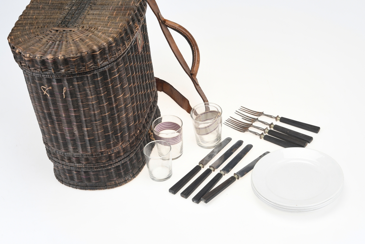 En piknikkurv med følgende innhold: 1 matboks, 2 større vannglass, 2 mindre vannglass, 4 hvite tallerkener, 4 kniver, 4 gafler og 1 beholder for salt/pepper. Kurven er flettet av pil eller lignende tre og er i ulik nyanser av brunt. Kurven er todelt, og i den nederste delen er det plass til matboksen, mens i den øverste delen det øvrige av piknikkutstyr. Beggel delene er foret med tesktil som er behandlet med granitol og deretter farget grønt. Langs kanten oppe (i begge delene) er det en svart tungekant av skinn. I den øverste delen av kurven er det festet strikk i lokket og i sidene i kurven for å feste utstyr. Det er også laget fire rom for å sette piknikkutstyr i. Rommene er laget av tekstil behandlet med granitol og farget grønn. Utvendig er det lærreimer som holder under- og overdel sammen samt fester lokket. De har spenner av messing og er dermed justerbare. I front er det et lite beslag med lås i messing. I den nederste deln av kurven er det plass til en oval matboks av blikk. Den har runde håndtak i hver side. Lokket er todelt og hengslet på midten slikk at lokkene åpnes mot midten. Inni boksen er det en skillevegg. Resten av utstyret er i "hovedkurven". Det er fire (vann)glass der i to forskjellige størrelser.  I de to største glassene er det en 1,5cm bred strikk som hindrer gnisning når de to mindre glassene er plassert i de to større. I tillegg er det fire hvite, enkle tallerkener. De har stempel på undersiden. Kurven innholder fire kniver og fire gafler. Knivene har avrundet blad av stål og svart trehåndtak. Gaflene har tre tinder av stål og svart trehåndtak. Til sist er det en beholder for salt og pepper. Den er av svart tre med dreid knopp på toppen. Skrur man av den øverste delen er det beholder for pepper. Skrur man av den nederste delen er det beholder for salt.