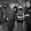 Christian Diors besök på NK, Nordiska kompaniet i Stockholm februari 1957. En grupp människor i varuhuset framför montrar. Kurt Jacobsson visar glasföremål.