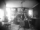 Interiör hos familjen Rythén. I centrum vid ett bord sitter A W och A M Rythén. Bakom dem står möjligen barnbarnet, senare fotografen, Anna-Lisa Rythén. Till vänster utmed väggen sitter dottern Anna Rythén.