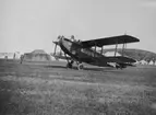 Brittiskt flygplan De Havilland DH. 34 märkt G-EBBQ (prototyp) står på ett fält under Internationella luftfartsutställningen i Göteborg (ILUG) 1923. Förare på plats i planet. Tälthangarer i bakgrunden.