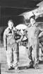 Gruppfoto av två militära flygare i flygarklädsel ståendes framför flygplan i hangar, 1920-tal.