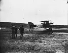 Flygplan FVM S 18 nr 982 står bock på ett fält. 1919. Flygförare i sitsen och tekniker i arbete vid vingarna.
