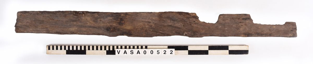 Bräda med två urtag och fem genomträngande spikhål, varav ett löper diagonalt genom ett hörn.
Mycket vittrad mot en kant. Järnlämningar kring spikhål. Avbruten i ett urtag.