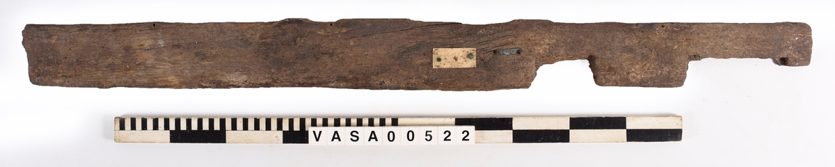 Bräda med två urtag och fem genomträngande spikhål, varav ett löper diagonalt genom ett hörn.
Mycket vittrad mot en kant. Järnlämningar kring spikhål. Avbruten i ett urtag.