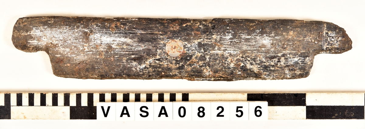 Odekorerad rektangulär bräda med mycket järn impregnering. 2 synliga spikhål, 4 kvister. 
Avbruten längs båda långkanterna. Ställen med järn/ saltskada på ena långkanten, rund spikhål, och på änden med fyndnummer. Fuktig och vittrade på alla ytor. Kraftig saltskada.
Tjock lager peg. Ett spår från konservering.