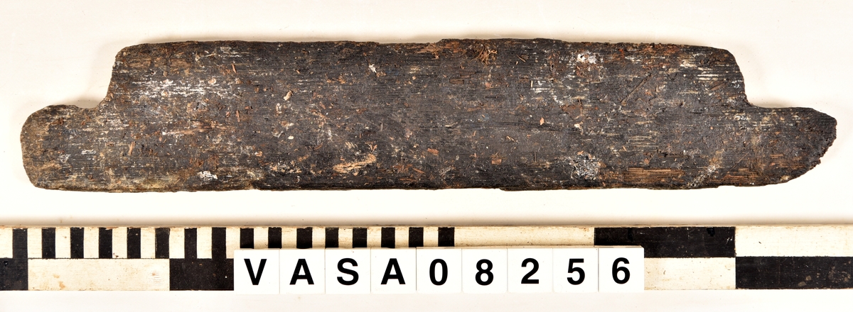 Odekorerad rektangulär bräda med mycket järn impregnering. 2 synliga spikhål, 4 kvister. 
Avbruten längs båda långkanterna. Ställen med järn/ saltskada på ena långkanten, rund spikhål, och på änden med fyndnummer. Fuktig och vittrade på alla ytor. Kraftig saltskada.
Tjock lager peg. Ett spår från konservering.