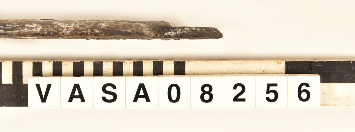 Odekorerad rektangulär bräda med mycket järn impregnering. 2 synliga spikhål, 4 kvister. 
Avbruten längs båda långkanterna. Ställen med järn/ saltskada på ena långkanten, rund spikhål, och på änden med fyndnummer. Fuktig och vittrade på alla ytor. Kraftig saltskada.
Tjock lager peg. Ett spår från konservering.